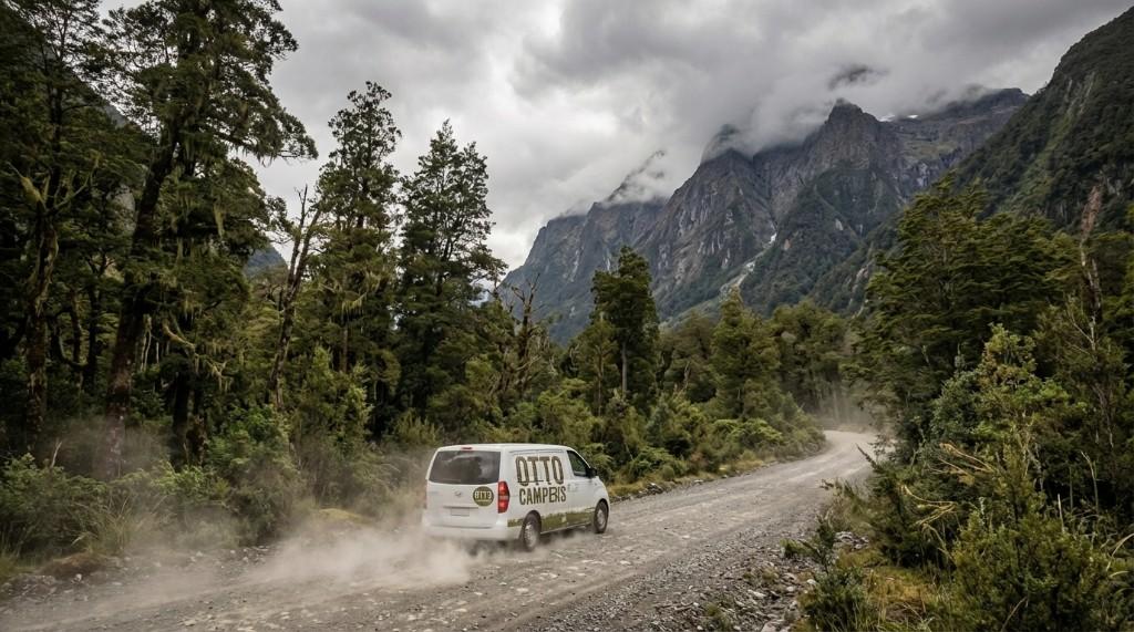 A white Otto Campers van on a dusty gravel road through forest with misty mountains ahead